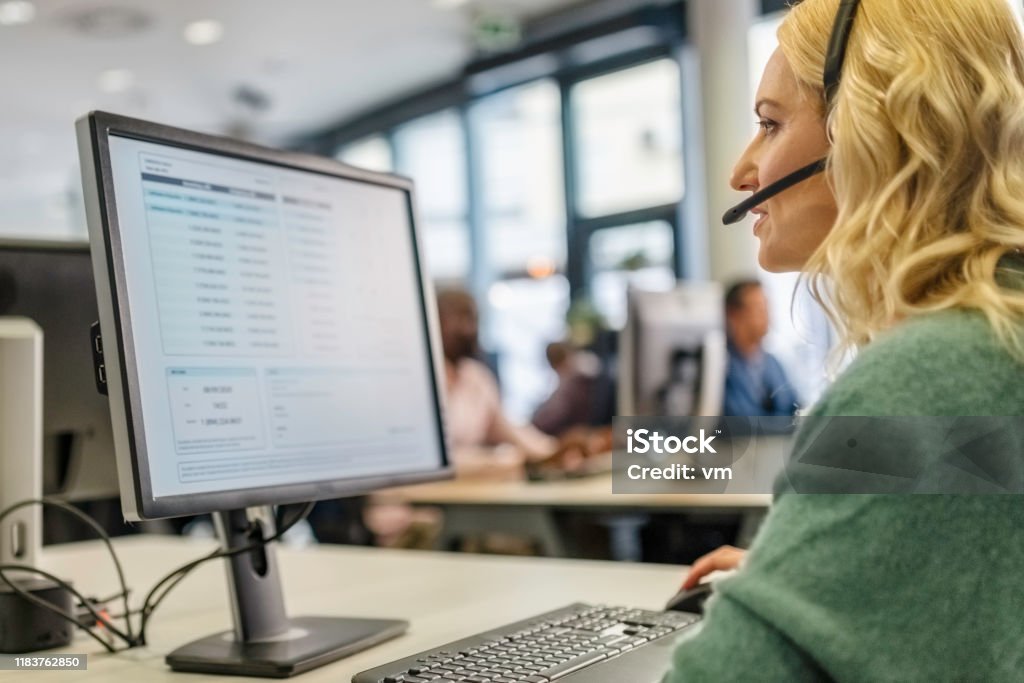 Woman working in a call centre Blonde woman with a headset with a microphone using a computer while talking to a customer in a call center. Customer Service Representative Stock Photo