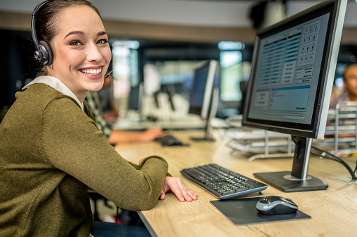 Portrait of a young woman in a call center looking at camera while leaning on a desk with a computer.