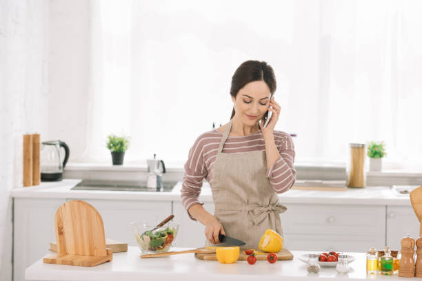 istock smiling woman in apron cutting bell pepper while standing near kitchen table with vegetables and talking on smartphone 1183763409