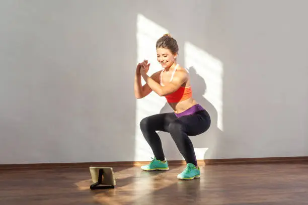 Photo of Positive sportive woman with bun hairstyle and in tight sportswear doing squatting. indoor studio shot illuminated by sunlight from window