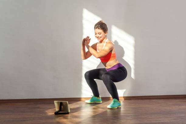 mulher desportiva positiva com penteado de coque e em sportswear apertado fazendo agachamento. estúdio interno disparado iluminado pela luz solar do indicador - crouching - fotografias e filmes do acervo