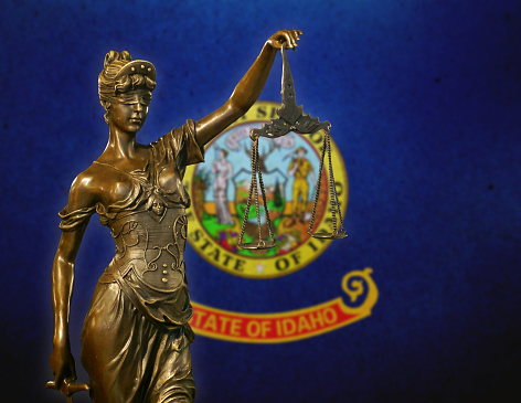 Close-up of a small bronze statuette of Lady Justice before a flag of Idaho.