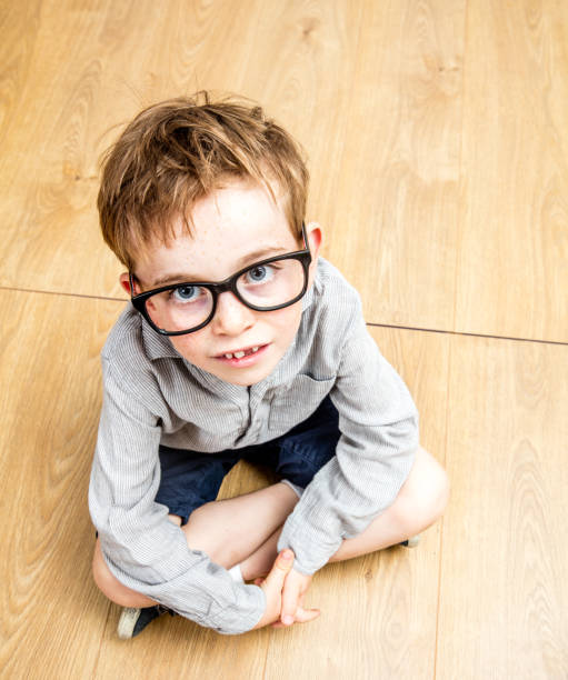 smart schoolboy with oversized eyeglasses looking up - high angle view smart schoolboy with oversized eyeglasses for genius nerdy look looking up, feeling inferiority, innocence or denigration, seated, crossed legs - high angle view denigrate stock pictures, royalty-free photos & images
