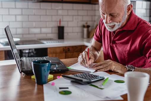 Senior man is sitting at the table, using a lpatop and planning home budget and writing something on the paper