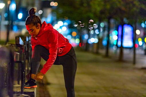 Young woman tying her shoelaces before running on city street at night. Young beautiful athletic girl standing on sidewalk and tying laces during training outdoors at night.