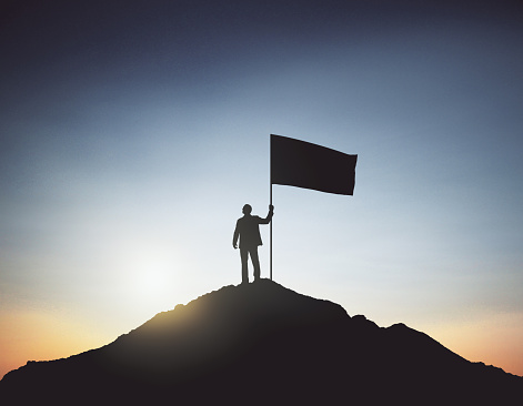 Backlit businessman with flag standing on mountain top. Sky background. Leadership and winner concept