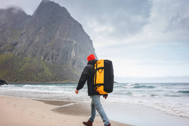 samotny odważny turysta z plecakiem wędrówki po piasku morskim wśród gór. podróżnik spacerujący po plaży oceanicznej przed wielką górską skałą. przygoda podróżnicza stylu życia - rock norway courage mountain zdjęcia i obrazy z banku zdjęć