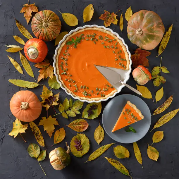 Flat lay of pumpkin pie with organic pumpkins and autumn leaves on black background. Thanksgiving concept overhead view