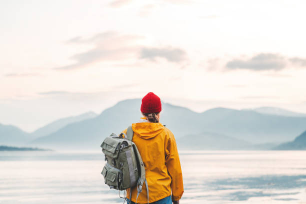 back view of male tourist with rucksack standing on coast in front of great mountain while journey.  man traveler wearing yellow jacket with backpack explore scandinavia nature. wanderlust outdoor - rock norway courage mountain imagens e fotografias de stock