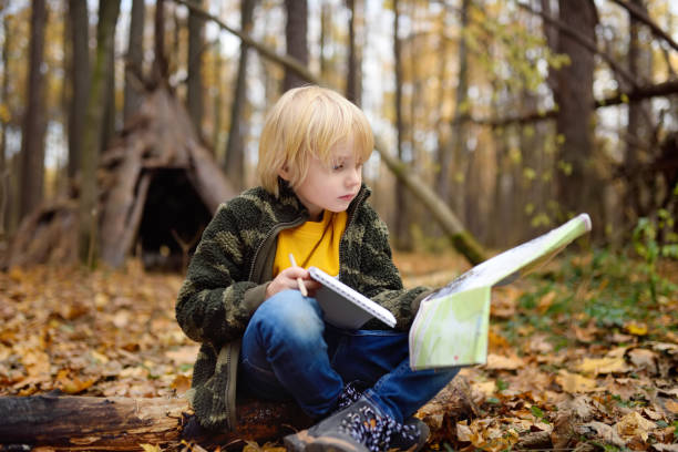 istock Little boy scout is orienteering in forest. Child is sitting on fallen tree and looking on map on background of teepee hut. 1183813047