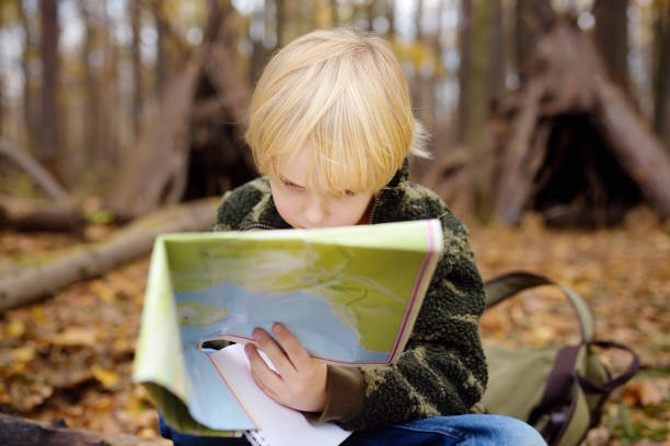 istock Little boy scout is orienteering in forest. Child is sitting on fallen tree and looking on map on background of teepee hut. 1183813103