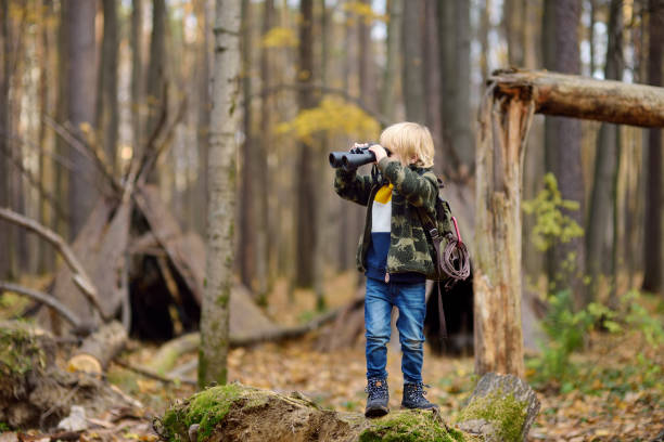 istock Little boy scout with binoculars during hiking in autumn forest. Child is looking through a binoculars. Behind the child is teepee hut. 1183813507