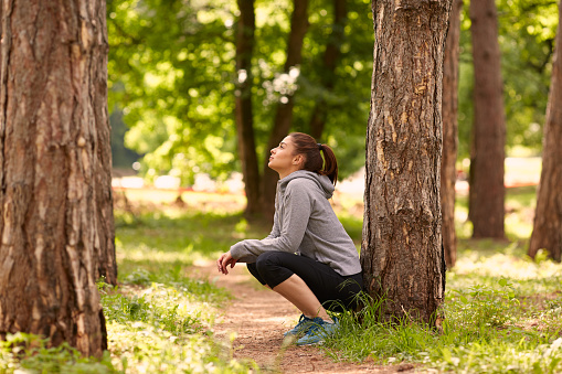 Side view of beautiful caucasian brunette with ponytail and in sportswear crouching in woods and resting after running.