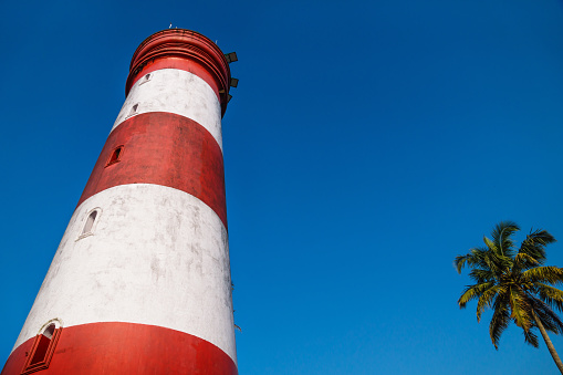 Low angle view of Alleppey Lighthouse with red and white stripes with palm tree on cloudless blue sky, Aleppuzha, Kerala, India