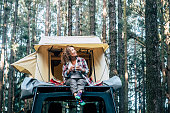 Happy cheerful beautiful people caucasian adult woman sit down on the roof of the car with tent in background and forest around - concept of travel and enjoy nature
