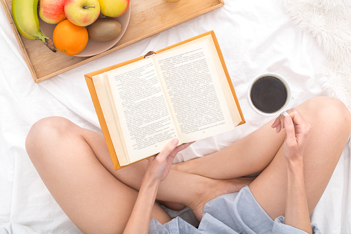 Young woman sitting on bed top view reading novel book drinking americano coffee