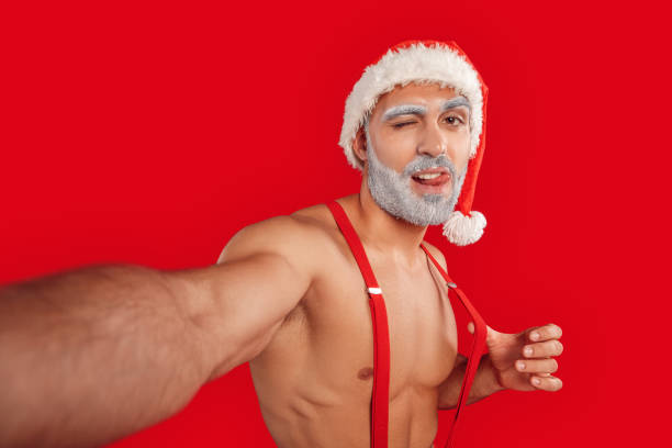 weihnachten freestyle. junge bärtige weihnachtsmann nackten muskulösen oberkörper in hut stehen isoliert auf rot machen selfie auf handy nahaufnahme zeigt zunge augenzwinkernd verspielt - sensuality men shirtless studio shot stock-fotos und bilder