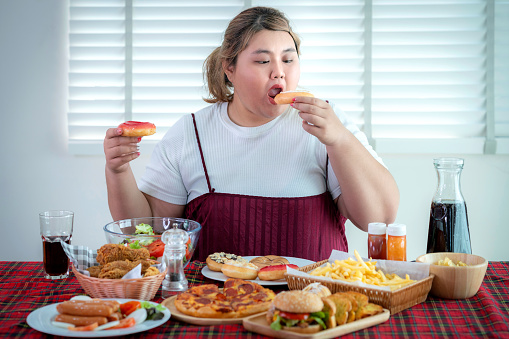 Asian fat girl hungry and eat a junk food on the table, this image can use for pizza, hot dog, sausage, doughnut, hamburger, potatoes, fried, french fries and fat concept