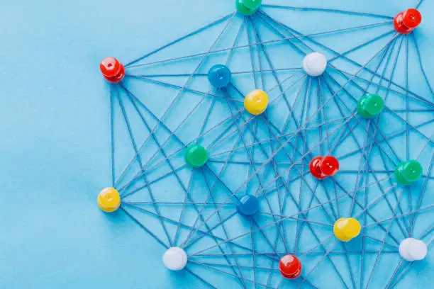 Photo of Small network of pins (Thumbtack)and string, An arrangement of colorful pins linked together with string on a pale blue background suggesting a network of connections.