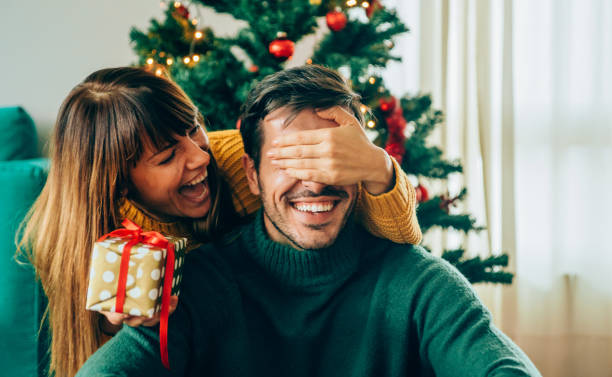 Romantic young couple exchanging Christmas gifts Beautiful young woman is standing behind her boyfriend with a gift in one hand and covered his eyes with the other hand. christmas present stock pictures, royalty-free photos & images
