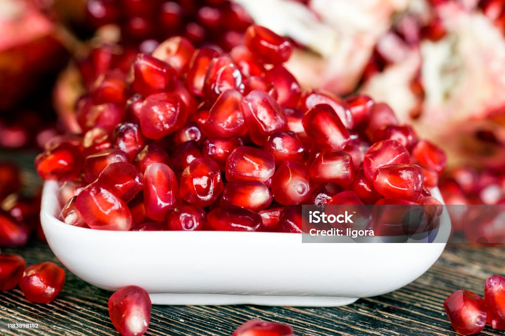 ripe pomegranate tasty sweet grains of ripe pomegranate on a wooden table, close-up of wholesome berries while breaking a whole fruit , small grains in a white bowl Acid Stock Photo