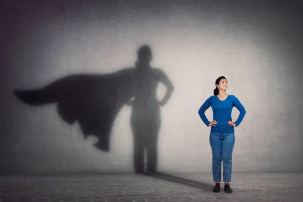 Photo of Brave woman keeps arms on hips, smiling confident, casting a superhero with cape shadow on the wall. Ambition and business success concept. Leadership hero power, motivation and inner strength symbol.