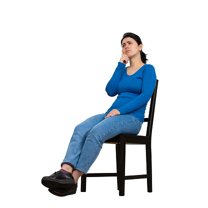 Side view full length portrait of casual young woman seated on a chair keeps hand under chin thoughtful looking away isolated over white background with copy space.
