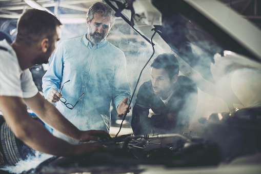 Mid adult customer and mechanics feeling frustrated while the car is smoking in a repair shop.