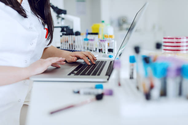 asistente de laboratorio caucásica dedicada sentada en el escritorio y usando computadora portátil para la entrada de datos. en el escritorio hay tubos de ensayo con sangre y platos de petri. - laboratory assistant healthcare and medicine chemistry fotografías e imágenes de stock