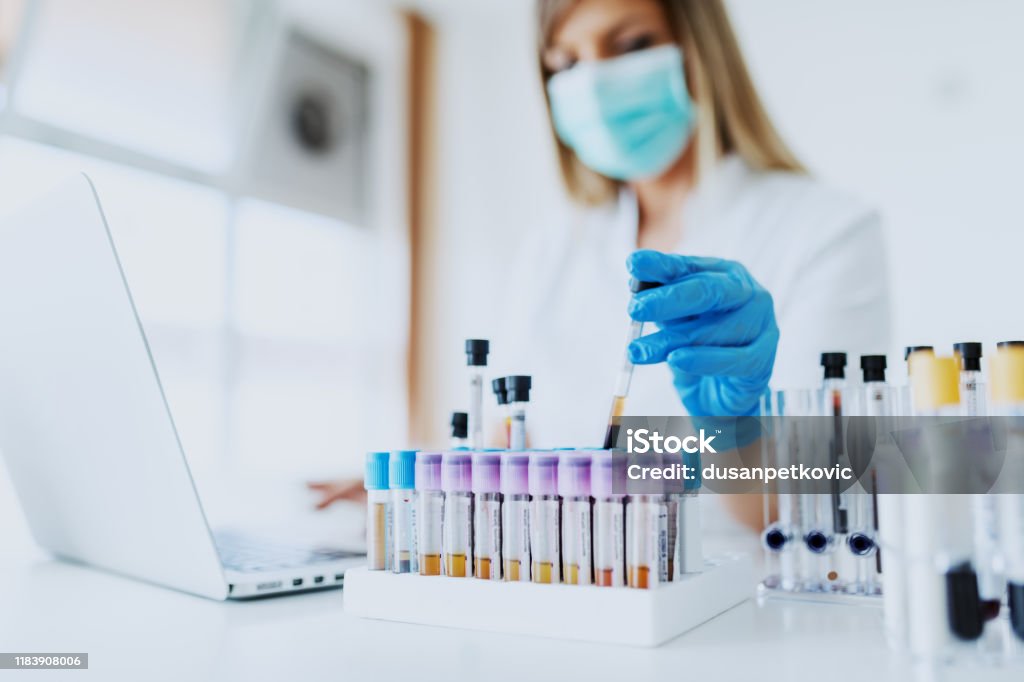 Close up of lab assistant in uniform, with mask and rubber gloves holding test tube with blood sample while sitting on chair and typing on laptop. Selective focus on test tubes. Laboratory Stock Photo