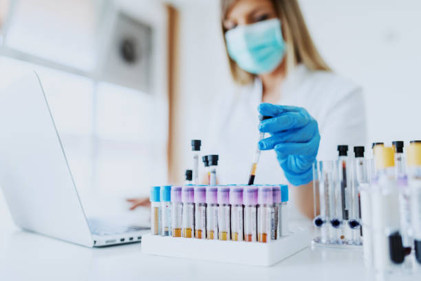 Close up of lab assistant in uniform, with mask and rubber gloves holding test tube with blood sample while sitting on chair and typing on laptop. Selective focus on test tubes. Close up of lab assistant in uniform, with mask and rubber gloves holding test tube with blood sample while sitting on chair and typing on laptop. Selective focus on test tubes. hiv stock pictures, royalty-free photos & images