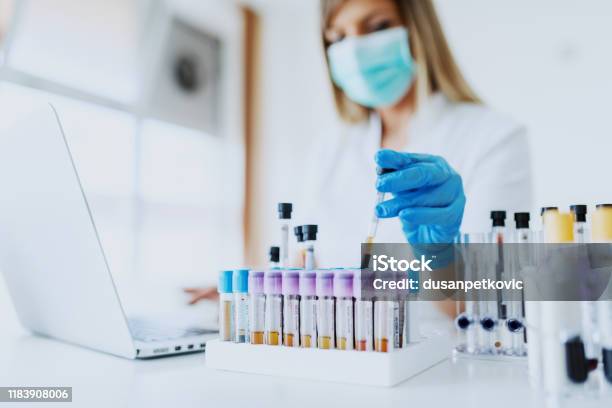 Close Up Of Lab Assistant In Uniform With Mask And Rubber Gloves Holding Test Tube With Blood Sample While Sitting On Chair And Typing On Laptop Selective Focus On Test Tubes Stock Photo - Download Image Now