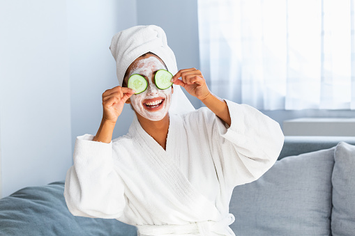 Woman with facial mask and cucumber slices in her hands. Beautiful young woman with facial mask on her face holding slices of fresh cucumber. Young woman with clay facial mask holding cucumber slices