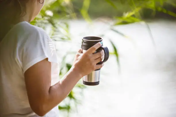 Cropped picture of mid adult woman standing by river, holding reusable cup.
