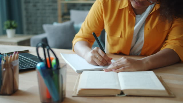 Close-up of female hand writing in notebook while woman reading book at table