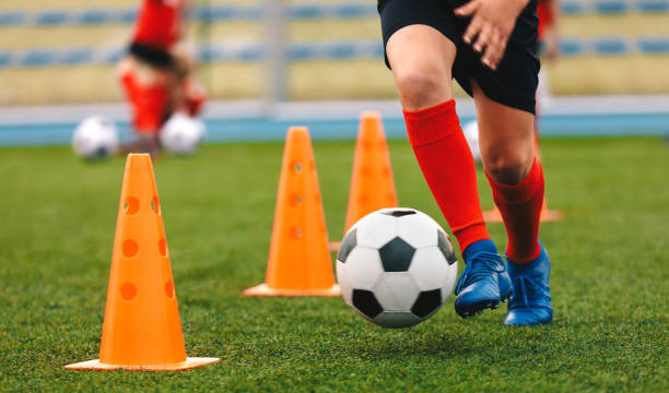 calciatore che dribbla palla in allenamento tra coni arancioni. giovane calciatore con tacchette blu sportive e calzini rossi - drill foto e immagini stock