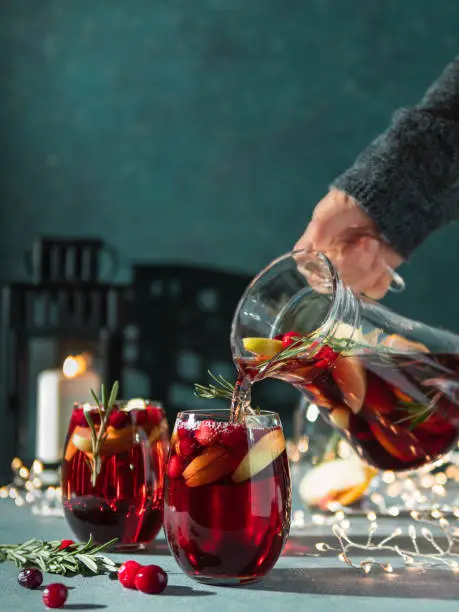 Female hand pours winter sangria in glasses with fruit slice, cranberry and rosemary. Dark christmas holiday background with candle, decoration lighting chain. Copy space for text or design. Vertical
