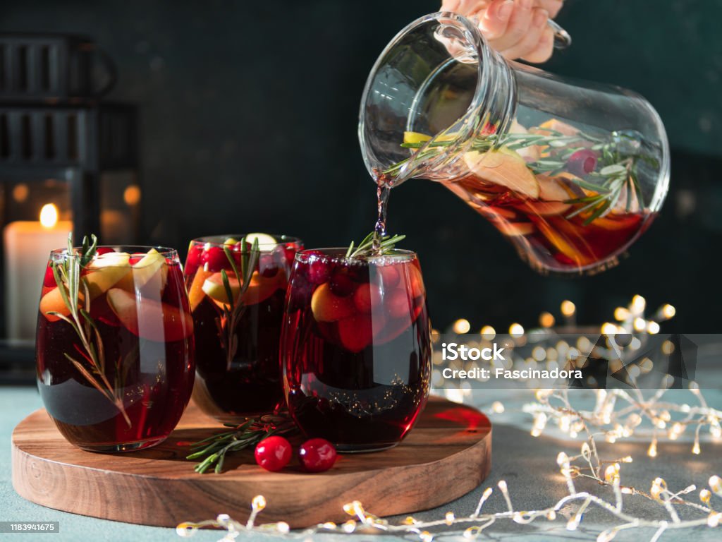 Winter sangria pouring in glasses, christmas table Female hand pours winter sangria in glasses with fruit slice, cranberry and rosemary. Dark christmas holiday background with candle, decoration lighting chain. Sangria Stock Photo