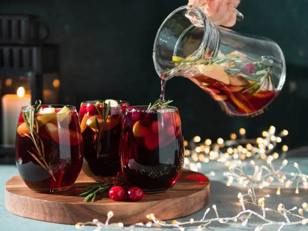 Female hand pours winter sangria in glasses with fruit slice, cranberry and rosemary. Dark christmas holiday background with candle, decoration lighting chain.