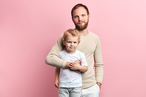 father and son looking at the camera while standing , happy fatherhood, parenthood. isolated pink background