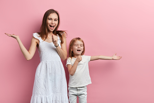 Excited cheerful happy mother and little daughter enjoying singing, close up portrait, isolated pink background, studio shot, entertainment, concert, performane