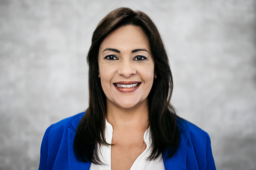 Indoor close-up of cheerful mature Hispanic businesswoman wearing v-neck blouse and royal blue blazer against gray background.
