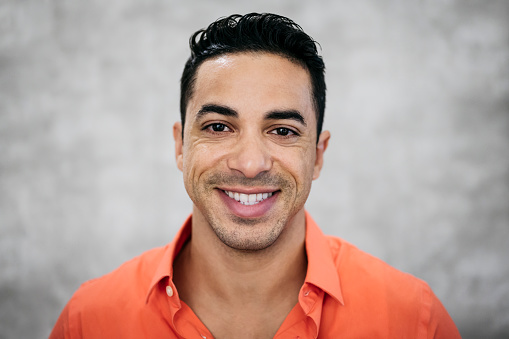Close-up headshot of mid adult Latin American businessman with positive attitude wearing open collar button up orange shirt against gray background.