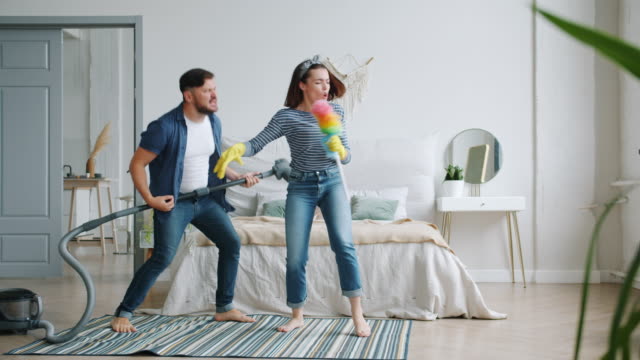 Woman and man singing in duster playing the guitar on vacuum cleaner in bedroom