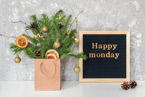 Photo of Happy Monday text on black letter board and festive bouquet of fir branches with christmas decor in craft package on table. Template for postcard, greeting card Concept Hello winter Monday