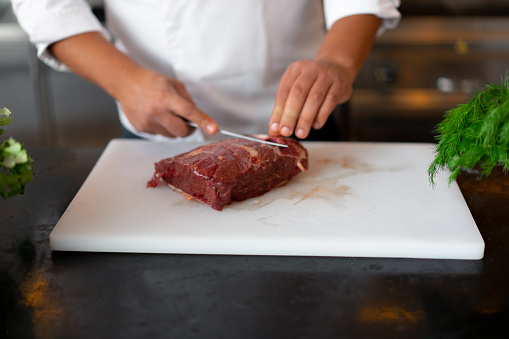 unrecognizable young African chef standing in professional kitchen in restaurant preparing a meal of meat and cheese vegetables. Portrait of man in cook uniform Cuts meat with a metal knife.