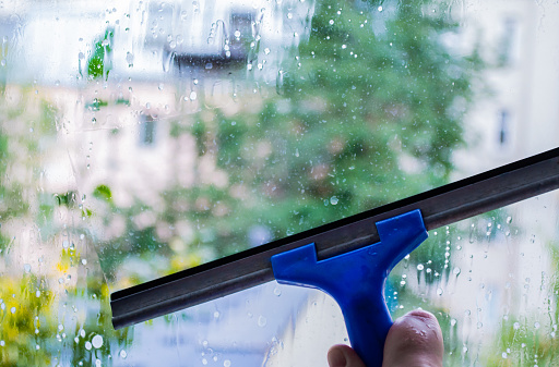 Window pane cleaning, housework with special squeegee. Outside the window blurred street, houses, greenery. The concept of cleanliness, cleaning, spring