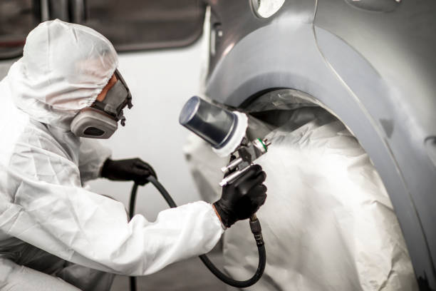 Painting technician applying a coat of paint on a car wheel panel segment in a body shop painting chamber Car body shop technician in full coveralls and a breathing mask applying a coat of paint on a car part in a painting booth. door panel stock pictures, royalty-free photos & images