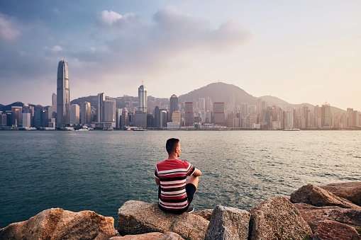 Young man sitting on stones against Hong Kong cityscape with skyscrapers at colorful sunset.