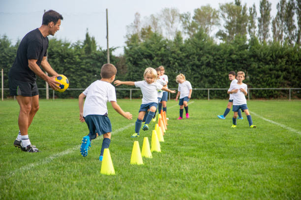 Young Footballers Practicing Running Drills During Practice Fit male coach in early 40s guiding young boy and girl Spanish footballers through practice drills around yellow traffic cones. sports training drill stock pictures, royalty-free photos & images